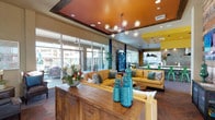 This dynamic single-floor community clubhouse is designed with a bold, eclectic aesthetic that balances energy and comfort throughout its interconnected spaces. The main lounge area immediately commands attention with a striking burnt-orange tray ceiling, exposed rustic wood accents, and a sculptural globe chandelier. Plush golden-yellow sofas and teal decorative pieces create a lively yet inviting atmosphere, while floor-to-ceiling glass walls flood the space with abundant natural light and provide direct views to the outdoor patio areas.

A secondary seating nook offers a more intimate retreat, featuring leather armchairs, curated abstract artwork, and candlelit side tables framed by large windows overlooking an exterior terrace. Nearby, a casual dining area with bold red metal chairs and industrial bistro tables opens directly to the outdoors via a full-panel glass garage door, blurring the boundary between inside and out.

The fully equipped kitchen is a standout feature, showcasing vivid yellow ceilings, a dramatic metallic hexagonal tile backsplash, dark cabinetry, stainless steel appliances, and a generous marble-topped island with vibrant green seating. A dedicated game area with an arcade machine adds a playful element, rounding out this well-appointed, amenity-rich gathering space designed for socializing and community engagement.