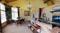 This single-story space makes a striking impression with its soaring vaulted ceilings accented by exposed wooden beams, creating an open and airy atmosphere. The warm golden-yellow walls are complemented by rich dark red architectural details, establishing a Mediterranean-inspired aesthetic throughout.

The main living area is anchored by a dramatic, organically shaped stone fireplace with ornate decorative metalwork, serving as a compelling focal point. A wrought-iron chandelier with candelabra-style lights hangs gracefully from the high ceiling, casting warm, ambient light across the room. Wall sconces further enhance the inviting atmosphere.

The space is furnished with a plush, neutral-toned sofa paired with a glass and wood coffee table featuring decorative ironwork. A patterned area rug defines the seating area. Adjacent to the main seating zone is a dining nook with ornate wooden chairs and a marble-top table, positioned near large windows that invite natural light into the space.

A gilded framed mirror adorns one wall, adding depth and elegance. The combination of textured stone, warm carpeting, and carefully curated furnishings gives the space a refined yet welcoming character.