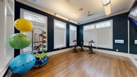This fitness studio is thoughtfully designed to support a variety of workout routines within a single, open space. The room is defined by bold navy blue walls accented with crisp white crown molding and baseboards, creating a polished yet energizing atmosphere. Warm hardwood-style flooring runs throughout, providing a durable and visually appealing surface suited for both cardio and floor-based exercises.

Natural light enters through multiple large windows fitted with white horizontal blinds, each topped with transom panes that draw the eye upward and enhance the sense of openness. Overhead, linear fluorescent lighting and a ceiling fan with a light fixture provide ample illumination and airflow during workouts.

A row of stationary spin bikes is positioned along the windowed wall, ideal for cycling sessions. Across the room, a large wall mirror reflects the space and aids in form correction, while a wall-mounted flat-screen television offers entertainment or guided workout programming. A dumbbell rack holds a graduated set of free weights, and a dedicated equipment stand organizes resistance bands, medicine balls, and stability balls. Yoga mats and foam rollers are neatly stored nearby. A glass interior window and door connect this studio to an adjacent fitness area, extending the overall functionality of the facility.