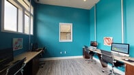 Central Cortex, located in Oxford, Mississippi, is a dynamic and visually striking workspace designed to foster productivity and creativity. The single-floor layout is unified by bold teal-painted walls that give the space a modern, energetic ambiance. Light wood-style flooring runs throughout, providing a clean contrast to the vivid wall color and contributing to a bright, open feel.

The workspace is equipped with multiple workstation areas, each featuring long, shared desks paired with ergonomic rolling chairs and desktop computers, accommodating several users simultaneously. Workstations are thoughtfully arranged along the walls, maximizing usable floor space while encouraging a collaborative environment.

Natural light is a defining feature of the space, with large windows along one wall allowing generous daylight to fill the room. Additional interior windows offer sightlines between areas, enhancing the sense of openness. White trim and clean architectural lines complement the vivid wall color for a polished, professional look.

Colorful abstract artwork is displayed throughout, adding personality and an artistic dimension to the workspace. Tall ceilings with elongated vertical architectural elements further amplify the sense of height and spaciousness. A printer station is also available, rounding out the functional amenities. Overall, Central Cortex delivers a well-equipped, visually engaging environment well-suited for collaborative or individual work.