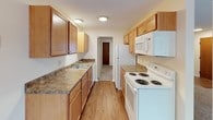 This single-floor unit offers a practical and comfortable layout designed for everyday living. The entry opens into a spacious living area finished with neutral wall-to-wall carpeting and cream-toned walls, creating a clean and versatile backdrop. A wall-mounted air conditioning unit and baseboard heating system serve the space, while a large window allows natural light to filter in, offering views of surrounding greenery.

The galley-style kitchen is efficiently organized, featuring warm-toned wood cabinetry both above and below granite-look laminate countertops. A double-basin sink, electric coil range, over-the-range microwave, and full-size refrigerator are all included. Light wood-style flooring runs throughout the kitchen, adding warmth and contrast to the adjoining carpeted areas.

A partial wall separates the living space from the sleeping area, with a hallway leading to what appears to be a closet or additional storage. The bedroom is well-proportioned, featuring two windows with blinds that provide ample natural light, along with baseboard heating. The textured ceiling is consistent throughout, and the overall neutral palette of the unit offers a move-in ready foundation easily adaptable to various furnishing styles.