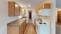 This single-floor unit offers a practical and comfortable layout designed for everyday living. The entry opens into a spacious living area finished with neutral wall-to-wall carpeting and cream-toned walls, creating a clean and versatile backdrop. A wall-mounted air conditioning unit and baseboard heating system serve the space, while a large window allows natural light to filter in, offering views of surrounding greenery.

The galley-style kitchen is efficiently organized, featuring warm-toned wood cabinetry both above and below granite-look laminate countertops. A double-basin sink, electric coil range, over-the-range microwave, and full-size refrigerator are all included. Light wood-style flooring runs throughout the kitchen, adding warmth and contrast to the adjoining carpeted areas.

A partial wall separates the living space from the sleeping area, with a hallway leading to what appears to be a closet or additional storage. The bedroom is well-proportioned, featuring two windows with blinds that provide ample natural light, along with baseboard heating. The textured ceiling is consistent throughout, and the overall neutral palette of the unit offers a move-in ready foundation easily adaptable to various furnishing styles.
