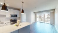 This single-floor residence at 230 E River Pkwy in Champlin, Minnesota, showcases a modern design with clean lines and a neutral color palette throughout. The open-concept layout connects the kitchen and living area seamlessly, creating a bright, cohesive space enhanced by large sliding glass doors that lead to a private balcony, offering outdoor views of the surrounding building complex.

The kitchen is well-equipped with white shaker-style cabinetry extending to the ceiling, stainless steel appliances including a refrigerator, electric range, microwave, and dishwasher. A quartz-topped island with a pendant light serves as a central focal point, complemented by a herringbone tile backsplash and warm wood-tone flooring.

Bedrooms feature plush carpeting, generous closet space, and large windows that bring in natural light. The primary bathroom includes a wide vanity with white cabinetry, a large mirror, and a glass-enclosed shower, while a secondary bathroom offers a tub-shower combination with brushed nickel fixtures. Wood-look flooring carries through the bathrooms, reinforcing a consistent, contemporary aesthetic. The building exterior features a modern façade with glass balcony railings and warm wood-panel accents.