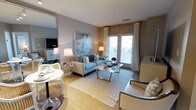 This well-appointed single-floor residence offers a thoughtfully designed open-concept layout that seamlessly blends the living and dining areas into one cohesive, welcoming space. Light wood flooring runs throughout, lending warmth and a sense of continuity to the interior. The living area is anchored by a soft blue sofa accented with teal throw pillows, paired with a glass-topped coffee table featuring gold-toned legs, and a complementary neutral armchair — all arranged atop a patterned area rug. A large abstract artwork in muted blues and purples serves as a striking focal point on the wall above the sofa. Elegant floor lamps provide ambient lighting, enhancing the room's refined atmosphere. French doors with white curtains and horizontal blinds flood the space with natural light while offering access to the outdoors. Adjacent to the living area, a compact dining nook features a round white table with modern tulip-style chairs, ideal for intimate gatherings. A wall-mounted television and mirrored panel further enhance the sense of space and depth. The overall palette of soft grays, warm neutrals, and cool blues creates a calm, contemporary ambiance that is both functional and visually inviting.