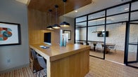 This single-floor space combines modern design with practical functionality, creating a polished and professional environment. The main workstation area is anchored by a generous wood-topped counter with built-in desktop computers, making it well-suited for focused individual work. Overhead, a trio of black dome pendant lights hang from a wood-paneled ceiling, casting warm, directed light across the workspace and adding a refined industrial touch.

Adjacent to the workstation, a sleek black steel-framed glass partition wall separates a private conference or meeting room. This room features exposed white stone walls that introduce a rustic, textural contrast against the otherwise contemporary aesthetic. A dining-style meeting table surrounded by modern white chairs occupies the space, illuminated by a coordinating pendant light fixture.

The patterned carpet throughout ties the two zones together visually, while the transparent glass partition maintains an open, connected feel across the space. The overall ambiance is professional yet inviting, balancing warm wood tones, natural stone textures, and modern black metal accents to create a cohesive, well-appointed environment suited for both collaborative and individual use.