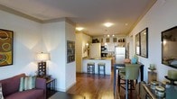 This single-floor residence offers an open-concept layout that seamlessly connects the living, dining, and kitchen areas into one cohesive and functional space. Rich hardwood flooring flows throughout, adding warmth and continuity to the interior.

The kitchen serves as a central hub, anchored by a peninsula with seating for two bar stools — ideal for casual dining or entertaining. It is equipped with stainless steel appliances, including a full-size refrigerator, and features white cabinetry that extends to the upper walls, providing generous storage. Track lighting and pendant fixtures illuminate the workspace, while decorative accents atop the upper cabinets lend a personalized touch.

The dining area is positioned adjacent to the kitchen, furnished with a high-top table and cushioned chairs, creating an inviting space for meals. A partial wall divides the dining and living zones, offering a subtle sense of separation without disrupting the open flow of the layout.

The living area is warmly lit with a table lamp and accented with framed wall art, contributing to the overall inviting ambiance. The space strikes a balance between comfort and style, with thoughtful details throughout that make this a well-rounded and livable single-floor home.