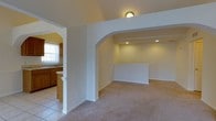 This single-floor residence offers a thoughtfully arranged layout that balances open living spaces with practical design. The main living area is finished with neutral-toned carpet and features a distinctive arched entryway that leads into a recessed lower section, adding architectural interest and a sense of depth to the space. Recessed lighting illuminates this area, while a small porthole-style accent light adds a unique decorative touch overhead.

The kitchen is a standout feature, designed in a galley style with rich warm-toned wood cabinetry lining both sides. White appliances, including a refrigerator and electric range with an overhead exhaust hood, are neatly integrated into the layout. Light tile flooring runs throughout the kitchen and into the adjoining dining area, providing a clean and cohesive look. A pendant light hangs above the dining space, which benefits from generous natural light streaming in through a large sliding glass door fitted with vertical blinds, offering views to the outdoors. The arched transition between the kitchen and dining area reinforces the architectural consistency found throughout the home. The overall ambiance is bright, neutral, and move-in ready.