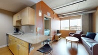 This single-floor urban unit blends modern industrial design with functional living spaces. The open-concept layout connects a well-appointed kitchen to a comfortable living area, creating a cohesive and efficient floor plan. The kitchen features light maple cabinetry, granite countertops, stainless steel appliances including a full-size refrigerator and range, a deep sink, and a breakfast bar with seating — ideal for casual dining. Pendant lighting and warm accent walls add character to the space.

The living area is furnished with leather seating, a round coffee table, and a television, all set against exposed concrete columns and visible ductwork that lend an industrial loft aesthetic. Large windows flood the space with natural light and frame views of the surrounding cityscape.

Two private bedrooms offer carpeted flooring, individual windows with urban views, and dedicated workspaces with desks and ergonomic chairs. Each room provides a quiet retreat with access to closet storage. The bathroom is finished with large-format tile flooring, a wood-toned vanity with ample storage, a wide mirror, and a walk-in shower area. Light maple wood doors and trim unify the design language throughout the unit, creating a cohesive and contemporary living environment.