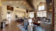 This expansive clubroom showcases a sophisticated design anchored by dramatic vaulted and cathedral ceilings that rise throughout the space, creating an open, airy atmosphere. Wood-look tile flooring flows continuously across all areas, unifying the various zones within the room. Natural light pours in through oversized windows and transom windows positioned high along the walls, complemented by sheer patterned drapery and recessed lighting overhead.

The main lounge area centers around a striking limestone-clad feature wall with a bold red-trimmed media niche housing dual flat-screen televisions, flanked by comfortable sofas and accent chairs arranged on a geometric area rug. Adjacent to the lounge, a wet bar with mosaic tile backsplash and bar-height seating provides an ideal gathering spot.

A dedicated game room offers a full-size pool table and shuffleboard, topped by a skylight that floods the area with daylight. The business center features built-in desks with computers and is complemented by nearby casual seating. A dining area with a round table and upholstered chairs sits near glass French doors that open to the outdoors. Curated abstract artwork and thoughtful accent furnishings complete the polished, welcoming ambiance throughout.