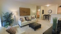 Located at 327 Guinevere On in Wilmington, North Carolina, this single-floor residence offers a well-designed, open layout that maximizes comfort and functionality. The living area serves as the heart of the home, featuring plush carpeting underfoot and neutral, warm-toned walls that create a welcoming ambiance. A large, eye-catching wooden wall art piece anchors the main seating area, complementing the light-colored sofa adorned with colorful accent pillows and a tufted leather ottoman that doubles as a centerpiece. Soft, ambient lighting from a floor lamp adds warmth to the space. The open floor plan transitions naturally into a dining area, where bar-height seating provides a casual yet defined space for meals. A hallway visible from the living room leads to additional areas of the home, including what appears to be a bathroom with warm cabinetry, suggesting thoughtful connectivity between spaces. The layout promotes an easy flow throughout the residence, making it both practical and inviting. Overall, the space strikes a balance between modern aesthetic touches and everyday livability, offering a comfortable and cohesive environment ideal for single-floor living.