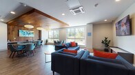 Located at 1401 King in Fairfield, Connecticut, this thoughtfully designed single-floor community amenity space blends modern aesthetics with functional versatility. The open-plan layout seamlessly connects a comfortable lounge area with a dining and social zone, creating a welcoming environment for gathering and relaxation.

The lounge area is anchored by deep navy blue sofas and armchairs accented with bold orange and red throw pillows, arranged around round wooden coffee tables atop a patterned area rug. A wall-mounted flat-screen television and a sleek media console complete this inviting sitting area. Glass-partitioned walls provide a view into an adjacent mailroom and corridor, maintaining a sense of openness throughout.

The dining area features dark rectangular tables paired with retro-style teal upholstered chairs, set beneath striking globe pendant lights that hang from a rich warm-wood ceiling accent. A vibrant abstract painting adorns the wood-paneled feature wall, adding a splash of color and artistic character to the space.

Large windows throughout flood the interior with natural light while offering pleasant views of the surrounding greenery and outdoor terrace. Durable wood-look flooring runs throughout the entire space, tying the zones together cohesively. A cozy built-in teal banquette nook provides an intimate seating option, while a small service counter adds further convenience. The overall ambiance is modern, bright, and welcoming.