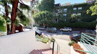 This residential building presents a distinctive Southern California architectural character, defined by its smooth stucco exterior finished in warm neutral tones with terracotta-tiled accents. The exterior immediately conveys a sense of established, well-tended living, with mature trees, palm trees, banana plants, lavender, and sculpted hedgerows framing the property and softening its street presence. A signature arched entryway adds architectural interest at the front, complemented by teal-painted metal railings and a secured gated entrance equipped with an intercom system, offering residents both style and security. Terracotta tile steps lead up to the entry level, reinforcing the warm, Mediterranean-influenced aesthetic. Walkways along the building's perimeter are accented with climbing vines and dense foliage, creating a lush, garden-like atmosphere. Balconies visible on the upper levels provide outdoor space with views of the tree-lined street and surrounding neighborhood. The building is situated on a quiet, greenery-rich street flanked by mature trees, offering a serene and shaded environment. The combination of gated access, verdant landscaping, outdoor lighting, and thoughtful architectural details reflects a property that balances comfort, character, and curb appeal in a desirable urban residential setting.