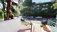 This residential building presents a distinctive Southern California architectural character, defined by its smooth stucco exterior finished in warm neutral tones with terracotta-tiled accents. The exterior immediately conveys a sense of established, well-tended living, with mature trees, palm trees, banana plants, lavender, and sculpted hedgerows framing the property and softening its street presence. A signature arched entryway adds architectural interest at the front, complemented by teal-painted metal railings and a secured gated entrance equipped with an intercom system, offering residents both style and security. Terracotta tile steps lead up to the entry level, reinforcing the warm, Mediterranean-influenced aesthetic. Walkways along the building's perimeter are accented with climbing vines and dense foliage, creating a lush, garden-like atmosphere. Balconies visible on the upper levels provide outdoor space with views of the tree-lined street and surrounding neighborhood. The building is situated on a quiet, greenery-rich street flanked by mature trees, offering a serene and shaded environment. The combination of gated access, verdant landscaping, outdoor lighting, and thoughtful architectural details reflects a property that balances comfort, character, and curb appeal in a desirable urban residential setting.