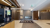 The Onyx at 1792 Pembina Highway in Winnipeg, Manitoba presents a thoughtfully designed lobby that makes a strong first impression. The entrance is anchored by a striking linear gas fireplace set into a grey tile feature wall, flanked by decorative wood slat columns and a built-in display of stacked firewood that adds a warm, organic contrast to the modern finishes. Large-format porcelain floor tiles in neutral tones extend throughout, contributing to the clean, contemporary aesthetic. A bold branded entry mat bearing "The Onyx" name reinforces the building's identity upon arrival. Adjacent to the main foyer, a dedicated mailroom area houses an extensive bank of individual resident mailboxes alongside a BlueBox smart parcel locker system, offering a convenient solution for package delivery. A concierge-style service counter with warm wood cabinetry and greenery provides a welcoming focal point near the elevator bank, which features teal and grey accent walls and carpeted corridors leading further into the building. Recessed lighting throughout ensures the space is well-illuminated, while wood-panelled ceiling details add warmth and depth. The overall ambiance is modern and inviting, balancing industrial-inspired materials with residential comfort.