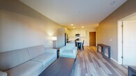 This single-floor unit offers a thoughtfully designed open-concept layout that seamlessly connects the living, dining, and kitchen areas. The living space is finished with wood-style plank flooring that extends throughout, lending a cohesive and contemporary feel. The kitchen is well-appointed with white shaker-style cabinetry, light countertops, and a full suite of stainless steel appliances, including a refrigerator, electric range, built-in microwave, and dishwasher. A kitchen island provides additional prep and dining space. Recessed lighting illuminates the kitchen and dining area, which accommodates a small dining table and chairs. The living area offers comfortable seating space with warm ambient lighting provided by a floor lamp. The bedroom features plush carpeting, a tray ceiling detail, and a window with an outdoor view, allowing natural light to fill the room. The bathroom is finished with the same wood-style flooring as the main living areas and includes a vanity with light countertops, gray cabinetry, a toilet, and a tub-shower combination. The overall ambiance of the unit is clean, modern, and functional.