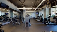 This fitness center offers a comprehensive workout environment designed to accommodate a wide range of exercise routines. The space is organized into distinct functional zones, allowing users to move seamlessly between cardio and strength training areas. The cardio section is lined with treadmills, elliptical machines, and stationary bikes, all positioned near large, multi-pane windows that invite natural light and provide views of the surrounding outdoor environment. A wall-mounted flat-screen television keeps users engaged during their sessions. The strength training area is outfitted with a robust selection of cable machines, weight benches, and a full dumbbell rack, all reflected in expansive floor-to-ceiling mirrors that enhance the sense of openness. An additional dedicated zone offers space for functional training with a power rack and resistance equipment. The flooring throughout consists of durable rubberized material suited for high-impact activity. The industrial-style ceiling features exposed ductwork, structural pipes, and a combination of linear LED and pendant lighting fixtures, lending the space a modern, urban aesthetic. Decorative wall art adds a refined touch, balancing the utilitarian design. The overall layout is open and spacious, facilitating a comfortable and efficient fitness experience.
