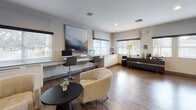 Located at 7960 Hampton Blvd in North Lauderdale, Florida, this single-story clubhouse offers a well-appointed community hub designed with a clean, contemporary aesthetic. The interior features warm hardwood-style flooring across the main areas, neutral walls, and recessed lighting that create a bright, welcoming atmosphere throughout.

The leasing and reception area is centrally anchored by a sleek white desk workstation with seating for residents and staff, complemented by easy access to private offices. These offices feature patterned carpeting, flush panel lighting, and dedicated desk setups framed by large windows that allow natural light to filter in. Contemporary abstract artwork adorns the walls, reinforcing the modern design language.

A resident lounge area provides comfortable modular seating, a wall-mounted flat-screen television, and stylish accent décor. Adjacent to the lounge, the business center offers a long built-in desk with multiple computer workstations, additional task seating, and ample cabinetry for storage. Windows throughout this area provide views of the surrounding greenery.

A transitional lounge space connects to a fitness area, visible through an interior window, housing cardio and strength equipment. Glass doors in this zone open directly to an outdoor patio, seamlessly connecting the interior to the exterior grounds.