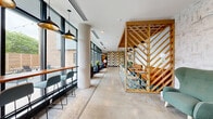 Located at 411 W Eighth St in Bloomington, Indiana, this thoughtfully designed hallway serves as a welcoming communal space that blends modern aesthetics with warm, eclectic character. Polished concrete floors run the length of the space, providing a sleek, durable foundation that contrasts beautifully with layered area rugs defining distinct seating zones. A dramatic chevron-patterned wood lattice partition anchors the center of the space, framing a lounge area furnished with rich caramel leather banquette seating, bistro-style chairs, and a vibrant patterned rug beneath. Floor-to-ceiling black-framed windows line one side of the hallway, flooding the interior with natural light and offering views of the surrounding Bloomington streetscape. A built-in counter with bar-height seating runs along the window wall, creating a functional workspace. Cylindrical white columns punctuate the corridor, adding architectural structure. At one end, a bold, whimsical wallcovering featuring illustrated cattle creates a striking focal point, complemented by textured white brick walls and framed artwork. Pendant lighting with black and gold-toned shades provides warm, layered illumination throughout. Potted plants and greenery add organic touches, completing this inviting, design-forward space.