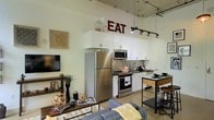 This single-floor space showcases a compelling blend of industrial and modern design elements. The open-concept layout connects the kitchen and living area in a fluid, functional arrangement. Soaring ceilings with exposed ductwork and piping overhead lend an urban, loft-like character to the space, while track lighting provides focused illumination throughout.

The kitchen is well-appointed with stainless steel appliances, including a full-size refrigerator, range, and microwave. White shaker-style cabinetry extends to the upper reaches of the tall walls, maximizing storage capacity. A subway tile backsplash adds a clean, classic detail behind the range and sink area. A compact dining table with metal bar stools is positioned adjacent to the kitchen, creating a defined dining zone without interrupting the open flow.

The living area features a plush sectional sofa and a wall-mounted flat-screen television, anchored by a striped area rug. A rustic wood console table serves as a decorative focal point, styled with books, plants, and decorative objects. The light-toned walls and concrete-style flooring unify the space, contributing to a bright, airy, and contemporary ambiance throughout.