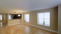 This single-floor space offers an open, airy layout highlighted by warm beige walls, clean white trim, and light-toned ceramic tile flooring throughout. The main living area is generously sized, benefiting from abundant natural light streaming through multiple large windows fitted with white blinds. The smooth, neutral-colored ceiling contributes to a clean, uncluttered aesthetic.

A pass-through opening connects the living area to the kitchen, which features a raised breakfast bar — ideal for casual dining or entertaining. Dark cabinetry, a tile backsplash with a decorative pattern, and darker appliances, including a refrigerator, give the kitchen a contrasting, contemporary feel against the lighter tones of the living space. A pendant light fixture hangs above the kitchen area, providing focused illumination and adding a subtle decorative touch.

The open layout between the living and kitchen areas promotes a seamless flow, making the space feel connected and functional. Large windows allow natural light to brighten the interior throughout the day, enhancing the warm, welcoming ambiance of the overall space.