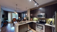 This one-bedroom, one-bathroom apartment at The Ashton in Spring, Texas offers a well-designed, single-story layout that balances modern aesthetics with functional living. The open-concept kitchen and living area serves as the heart of the home, where dark espresso cabinetry pairs with light granite countertops and a textured tile backsplash. Stainless steel appliances, including a range, microwave, and refrigerator, complement the kitchen's contemporary feel. A peninsula with an undermount sink and pendant lighting above creates a natural transition into the living space, which features hardwood-style flooring, a ceiling fan, and large windows with outdoor views that bring in generous natural light.

The living room offers a comfortable, open atmosphere with access to a private exterior door, adding convenience and connectivity to the outdoors. The bedroom is carpeted for warmth, featuring multiple windows, ceiling fan, and ample space for furnishings. The bathroom includes a soaking tub, tile surround shower, dark vanity cabinetry with a wide mirror, and hardwood-style flooring extending through the hallway. Track lighting and pendant fixtures throughout the unit add a polished, intentional design element to the overall ambiance.
