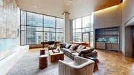 Located at 1699 Scotts Station Street in McLean, Virginia, this single-floor living space is defined by its dramatic architectural design and high-end finishes. The room commands immediate attention with soaring double-height ceilings clad in warm wood paneling, complemented by integrated cove lighting that casts a refined ambient glow throughout the space.

Floor-to-ceiling glass curtain walls span a generous portion of the room, flooding the interior with abundant natural light and offering expansive views of the surrounding McLean cityscape. The intricate parquet hardwood flooring adds warmth and texture underfoot, anchoring the contemporary aesthetic.

A striking gas fireplace serves as a dramatic focal point, encased in a floor-to-ceiling sculptural tile surround and framed by a glass front that showcases the flame. A wall-mounted television is positioned nearby, enhancing the entertainment functionality of the space. Opposite, a built-in wet bar features light wood cabinetry, open shelving, and a sink with an antique mirrored backsplash, offering a refined service area.

The living area is thoughtfully appointed with leather seating, a sculptural wood coffee table, and leather ottoman stools. A shuffleboard table near the windows adds a recreational dimension. The overall ambiance is one of sophisticated urban living, balancing warmth and modernity seamlessly.