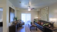 This single-floor space offers a welcoming and functional layout designed for comfortable everyday living. The living room serves as the centerpiece of the home, featuring warm, neutral-toned walls and a textured ceiling outfitted with a ceiling fan and integrated light fixture, providing both airflow and ambient illumination. Dark hardwood-style flooring grounds the space, adding a rich contrast to the lighter walls and ceiling. A large sliding glass door fitted with vertical blinds floods the room with natural light and provides direct access to an outdoor balcony or patio area, extending the living space beyond the interior. The room comfortably accommodates a full seating arrangement, including a dark leather sofa accented with colorful throw pillows and a patterned accent chair, anchored together by a woven area rug and a wooden coffee table. A dark wood media console along one wall adds storage and display functionality. Decorative touches, including potted greenery, table lamps, and wall art, contribute to the warm and inviting ambiance. Overall, the space balances practical design with thoughtful detail, creating a cozy yet visually appealing environment.