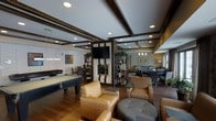 This well-appointed single-floor lounge space is designed for both recreation and relaxation, offering a range of amenities within an open, flowing layout. The room is anchored by a sleek billiards table positioned on rich hardwood flooring, making it an immediate focal point upon entry. A central fireplace with a clean, modern tile surround doubles as a media wall, with a large flat-screen television mounted above, creating a natural gathering hub within the space.

The ceiling features dark wood coffered beams that add architectural depth and visual interest, complemented by recessed lighting that provides consistent, warm illumination throughout. The lounge is organized into distinct yet connected zones: a casual seating area with woven armchairs and an ottoman, and a more formal dining or meeting area visible in the background with additional chairs and tables.

Open shelving units flank the fireplace, adding decorative flair while subtly defining the various areas. Neutral tones in the upholstery and walls are grounded by the dark wood accents, creating a cohesive, sophisticated ambiance. Natural light filters in from windows along the perimeter, enhancing the inviting atmosphere of this versatile communal space.