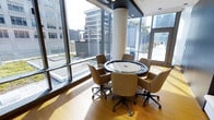 This contemporary commercial space is designed to foster collaboration and productivity in a bright, open environment. The standout feature is the expansive floor-to-ceiling glass curtain walls that flood the interior with abundant natural light while offering sweeping views of the surrounding urban skyline and outdoor terrace areas. The warm hardwood flooring contrasts beautifully with the dark ceiling accents, creating a polished yet inviting atmosphere. At the heart of the space sits a round conference table with a sleek, modern finish, surrounded by comfortable upholstered chairs on casters, accommodating small group meetings with ease. A structural column with a rounded profile adds an architectural element without disrupting the open feel of the room. The layout encourages open sightlines and connectivity to the outdoors, making the space feel considerably more expansive than its footprint suggests. Access to an adjacent outdoor terrace further enhances the space's versatility, providing an option for al fresco gatherings against a backdrop of city buildings. The overall ambiance is clean, professional, and contemporary, well-suited for focused meetings, collaborative sessions, or informal discussions in a refined urban setting.