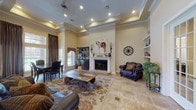 Located at 330 W Cheryl Dr in San Antonio, Texas, this single-story space showcases a thoughtfully designed interior with a warm, Western-inspired aesthetic throughout. The main living area is anchored by a tray ceiling with recessed lighting and a ceiling fan, lending an elevated sense of volume to the room. A white-mantled fireplace serves as a focal point, complemented by decorative wall sconces, rustic artwork, and a wagon wheel accent. Built-in shelving units flank the space, offering both display and storage functionality. Large-format travertine tile flooring flows consistently throughout, adding a cohesive, upscale feel. White decorative columns frame the transition into the kitchen, which features warm wood cabinetry, granite countertops, a stainless steel range hood, and ample counter space in a well-organized layout. An adjacent dining area benefits from soaring windows dressed with floor-length drapes, allowing generous natural light and views of the surrounding exterior. A set of glass-paned French doors connects the living room to an additional room, enhancing the open and connected flow of the space. The overall ambiance is inviting and well-appointed, blending classic architectural details with a distinctive Southwestern character.