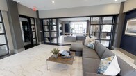 This single-floor space presents a sophisticated and contemporary lobby environment designed to impress and welcome guests. The interior is anchored by striking marble-style porcelain tile flooring that runs throughout, lending a sense of elegance and spaciousness to the open-plan layout.

A defining architectural feature is the use of bold, black steel-framed glass partitions that divide the space into distinct zones while maintaining visual connectivity and an airy, open feel. These floor-to-ceiling glass walls allow natural light to flow freely between areas, enhancing the bright and inviting atmosphere.

The main seating area is furnished with a generous grey sectional sofa accented by patterned throw pillows, paired with a rustic wood and metal coffee table topped with curated décor. A secondary lounge area, visible through the glass partitions, features additional soft seating and potted greenery, reinforcing the space's warm yet polished character.

Recessed ceiling lighting provides consistent, well-distributed illumination throughout. Natural stone accent walls add texture and depth, complementing the otherwise sleek, modern palette of grey, white, and black tones. The overall ambiance is one of refined, contemporary design — professional, comfortable, and thoughtfully appointed.