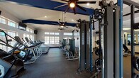 This fitness center offers a comprehensive workout environment spread across a single, open floor plan designed to accommodate a wide range of exercise routines. The space is architecturally distinguished by soaring vaulted ceilings accented with striking navy blue beams, complemented by large multi-pane windows that line the walls and flood the interior with natural light. Ceiling fans are strategically positioned throughout to maintain air circulation during intense workouts.

The cardio zone is well-appointed with a row of Matrix treadmills and upright exercise bikes, all positioned beneath wall-mounted flat-screen televisions for an engaging workout experience. The strength training area features a blue power rack with Olympic weight plates, a functional cable machine system, a multi-station cable tower, and a selection of selectorized machines targeting various muscle groups. An adjustable bench and a dumbbell rack with a full-length mirror wall round out the free weights section.

Rubber flooring covers the entire workout area, providing both durability and safety underfoot. A separate, lower-ceilinged annex houses additional free weight equipment, benches, medicine balls, and mirror walls, offering a quieter space for focused strength work. The fitness center is accessible via double glass-panel entry doors and includes additional interior doors leading to support rooms.