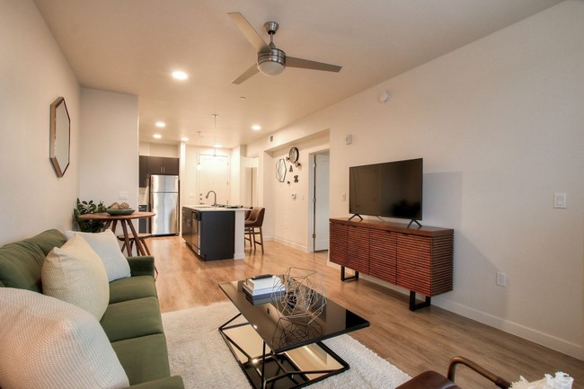 White living room with modern ceiling fan, view to kitchen with large island, and door to bedroom. - View 32 Apartments