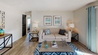 This single-floor residence centers around a well-appointed living room that sets a welcoming, coastal-inspired tone throughout the space. The living room features wood-style flooring that adds warmth and visual continuity, while neutral-toned walls provide a versatile backdrop for the light, airy décor. A textured popcorn ceiling adds character overhead, and a central HVAC vent ensures year-round comfort.

Natural light flows generously into the room through a sliding glass door dressed with teal curtains, offering views of the surrounding neighborhood and providing direct access to an outdoor area. The living space is comfortably proportioned, accommodating a tufted sofa, accent seating, side tables, and a wood-and-metal coffee table with ease.

Coastal-themed artwork adorns the walls, complementing the blue-and-white geometric area rug below. The open layout connects the living room to additional areas of the home, with a hallway visible leading toward what appears to be a bedroom, suggesting a practical and accessible floor plan. Overall, the space conveys a relaxed yet polished ambiance suitable for everyday living.