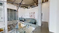 This business center showcases a striking industrial aesthetic defined by soaring ceilings with exposed ductwork, conduits, and concrete structural columns that lend the space a raw, urban character. The open-plan layout is organized across a single floor, with glass panel partitions creating visual separation between work zones while maintaining an airy, connected feel throughout.

The workspace features polished concrete floors that complement the industrial design language, while recessed and track lighting illuminate the interior effectively. A lounge or collaborative seating area is visible, furnished with contemporary gray sofas and equipped with workstations, offering a flexible environment for both focused work and group interaction.

Wall-mounted artwork adds a creative dimension to the space, reinforcing a professional yet dynamic atmosphere. Large windows along the perimeter allow natural light to filter in, softening the industrial elements. The combination of glass, concrete, steel, and white walls creates a clean, modern contrast that feels both functional and visually engaging — well-suited for a productive business environment.