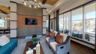 This well-appointed single-floor community clubhouse offers a thoughtfully designed layout that balances social functionality with refined aesthetics. The entrance welcomes visitors through glass double doors framed with transom windows, filling the reception area with natural light. Decorative wall-mounted sculptures and bold abstract artwork set an artistic tone from the moment you arrive.

The main lounge area is a standout feature, boasting elevated ceilings with exposed wood beams, an elegant chandelier, and a warm color palette of terracotta, gray, and cream. Floor-to-ceiling windows with draped curtains provide views of the outdoor surroundings, while plush seating arrangements and a wall-mounted flat-screen TV create an inviting environment for relaxation.

A dedicated billiards area adds a recreational dimension to the space, complete with a full-size pool table, another statement chandelier, and continued access to natural light through large windows. The kitchen is equally impressive, featuring dark cabinetry, a reflective tile backsplash, exposed beam ceilings, a central island with seating, and artisan patterned floor tiles that add a distinctive design element.

A casual dining nook with small round tables and warm-toned chairs is adorned with large-scale watercolor artwork, providing an intimate setting for conversation. Throughout, the hexagonal tile flooring and consistent design language tie the entire space together cohesively.