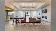 This single-floor residential clubhouse and leasing center presents a thoughtfully designed space that blends modern aesthetics with a warm, welcoming atmosphere. The grand entry lobby sets an immediate tone of sophistication, featuring large-format tile flooring, wall sconces, and a statement chandelier overhead. Glass double doors with stone exterior accents create a polished first impression.

The expansive main lounge area is anchored by two elegant crystal chandeliers suspended from a coffered ceiling. A mix of seating arrangements — including a large sectional sofa on a patterned area rug, a dedicated dining table with contemporary chairs, and a cozy fireplace lounge with armchairs — offers versatile social spaces. Multiple windows allow abundant natural light throughout.

A dedicated seating nook features steel-blue accent walls, exposed wood ceiling beams, and generous windows with views of the surrounding neighborhood, creating an intimate yet open retreat.

The fully equipped communal kitchen is outfitted with dark cabinetry, white quartz countertops, gold-framed open shelving with greenery, glass pendant lighting, and a wine refrigerator, providing a functional and stylish gathering space.

The leasing office area features a sleek dark desk with white quartz top, chandelier, and a private consultation window, rounding out this well-appointed community facility.