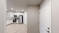 This single-floor space offers a clean, modern aesthetic defined by neutral gray and warm beige tones, light wood-style luxury vinyl plank flooring, and crisp white trim throughout. The kitchen is a standout feature, outfitted with white shaker-style cabinetry, granite-look countertops, and a full suite of stainless steel appliances including a refrigerator, electric range, and built-in microwave. Recessed lighting illuminates the kitchen area, complementing its bright, functional design. Adjacent to the kitchen, multiple doors provide access to additional spaces, suggesting ample storage or utility areas. The living area is open and versatile, featuring a thermostat, HVAC vents, and a dedicated return air vent, indicating a well-integrated climate control system. A bedroom with a ceiling fan and a window with blinds allows for natural light and airflow. Additional bedrooms are generously sized with neutral walls and the same consistent flooring. The bathroom is neatly finished with a white vanity, granite-look countertop, a tub-shower combo with tiled surround, and light wood-style flooring. Overall, the space presents a cohesive, move-in-ready interior with thoughtful, practical finishes throughout.