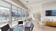 This contemporary amenity lounge is a thoughtfully designed single-floor communal space that seamlessly blends functionality with modern aesthetics. The layout encompasses three distinct zones, each contributing to an open, inviting atmosphere filled with abundant natural light.

The main lounge area is anchored by expansive floor-to-ceiling windows and glass doors that frame direct views of the outdoor pool courtyard. Elevated ceilings with structural beam detailing add architectural depth, while light hardwood flooring flows throughout, unifying the space. Roller shades provide adjustable light control without compromising the airy feel.

A full-service kitchen bar serves as a standout centerpiece, featuring a striking patterned mosaic tile backsplash, white cabinetry with gold hardware, quartz countertops, stainless steel appliances, and a generously sized island with bar seating illuminated by pendant lighting suspended from brass chain fixtures.

Separate seating and dining areas offer flexible gathering spaces, with multiple table-and-chair configurations suited for both casual lounging and more structured meetings. Large sliding glass doors provide direct outdoor access, reinforcing the seamless indoor-outdoor connection. The overall design palette — neutral tones, clean lines, and curated artwork — creates a polished yet welcoming communal environment.