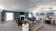 This well-appointed single-floor lounge space is designed with both comfort and style in mind. The room immediately draws the eye upward with its vaulted ceilings, which lend an open, airy quality to the interior. A classic white-framed fireplace serves as the focal point of the main seating area, flanked by dark accent walls with a sophisticated two-tone paint treatment — deep slate blue on the upper portion and crisp white wainscoting below — creating a refined, contemporary aesthetic.

The space is thoughtfully arranged into distinct zones. A generous main seating area features a plush taupe sectional sofa, sleek dark leather armchairs, and round white accent tables, all set atop soft wall-to-wall carpeting. Teal accent pillows and lush greenery add pops of color throughout. A wall-mounted flat-screen television above the fireplace enhances the room's entertainment functionality.

Toward the rear, a casual high-top dining area offers bar-height seating beside large windows that allow natural light to filter in. Pendant light fixtures provide warm, even illumination throughout. The overall ambiance is modern and inviting, balancing elegant design details with a relaxed, welcoming atmosphere suited for both quiet gatherings and social events.