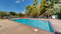 This outdoor pool area in Hattiesburg, Mississippi offers a well-maintained aquatic retreat designed for relaxation and recreation. The centerpiece is a large, rectangular swimming pool with clear blue water, bordered by a spacious concrete deck that provides plenty of room for lounging and movement. Numerous blue sling-style lounge chairs are arranged along multiple sides of the pool, giving users a variety of spots to relax and soak up the Mississippi sunshine.

The pool area is enclosed by a sturdy iron perimeter fence, enhancing both safety and privacy. Mature trees and ornamental plantings surround the space, creating a natural, shaded backdrop that softens the outdoor environment and provides welcome relief from the sun. A safety ring is visible along the fencing, reflecting attention to pool safety standards.

Access to the pool area is facilitated by steps with brick-trimmed borders, leading from an adjacent building with white vinyl siding and classic brick exterior construction. The overall setting benefits from abundant natural light and open sky views, contributing to a bright and inviting atmosphere. The combination of generous deck space, thoughtful landscaping, and practical amenities makes this pool area a comfortable and functional outdoor space.