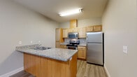 This single-floor residence offers a thoughtfully designed layout that balances open communal spaces with comfortable private areas. The open-concept kitchen is a central highlight, featuring light maple cabinetry, granite-look countertops, a peninsula with an integrated sink, and a full suite of stainless steel appliances including a refrigerator, range, and microwave. Durable wood-look laminate flooring runs throughout the main living areas, lending a clean, cohesive feel.

The living area is generously sized and filled with natural light from a sliding glass door that opens to an outdoor space, complemented by a ceiling fan overhead. Built-in wall shelving in the main area provides a functional and architectural detail. A dedicated hallway connects the living spaces to the bedrooms, which feature plush carpeting, ceiling fans, natural light from windows with blinds, and closet storage. The neutral, soft-toned walls throughout create a calm and versatile backdrop.

The bathroom is cleanly appointed with a full tub-shower combination, a wood-vanity with granite-style countertop, and wood-look flooring. Additional utility areas are also present, offering practical storage and flexible use. Overall, the space presents a clean, move-in-ready environment with consistent modern finishes.