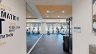 Located at 26 S York Rd in Hatboro, Pennsylvania, this well-appointed fitness center offers a comprehensive exercise environment across a single floor. The space is thoughtfully designed with a tray ceiling accented in warm tones, recessed lighting, and a ceiling fan that contribute to a clean, modern ambiance.

The main workout area is anchored by a row of cardio machines, including treadmills, ellipticals, and a stationary bike, positioned beneath large windows that allow generous natural light and views of the surrounding neighborhood. A wall-mounted flat-screen television is conveniently placed for entertainment during workouts.

A full-wall mirror spans one side of the room, visually expanding the space and providing useful feedback during exercise. Strength training is well-supported with a multi-functional power rack loaded with weight plates, a dedicated dumbbell rack, adjustable weight benches, and a medicine ball station. A Peloton-style interactive spin bike adds a connected fitness option.

The flooring throughout is a durable, cushioned dark rubber surface suited for high-impact activity. The open layout provides ample room for functional training and stretching. A set of double doors leads to additional storage or utility access. The overall design balances functionality and comfort, making it a capable and inviting fitness facility.