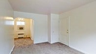 This single-floor space presents a clean, understated layout defined by neutral tones and practical design. The main living area is finished with wall-to-wall carpet in a muted gray-beige tone, creating a soft and grounded atmosphere throughout. White walls and standard-height ceilings contribute to a bright, straightforward interior.

A distinctive arched doorway connects the main living space to an adjacent room, adding a subtle architectural character to an otherwise simple floor plan. The adjoining room features a window that allows natural light to filter in, along with a wall-mounted air conditioning unit and a baseboard heating unit beneath, indicating year-round climate control capability. A chandelier fixture is partially visible through the archway, lending a modest decorative touch to that secondary space.

Back in the main area, two white-paneled doors are positioned side by side along one wall. The larger door, fitted with brass hardware, serves as the primary entry point, while the smaller door provides access to a closet. The overall design is functional and neutral, making the space highly adaptable to a variety of furnishing styles and uses.