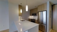 This single-floor space showcases a well-appointed modern kitchen designed with both functionality and aesthetics in mind. The kitchen is anchored by a spacious white waterfall island that doubles as a prep and casual dining area, complete with an undermount sink and a sleek arc faucet. Warm, flat-front wood cabinetry lines the walls, extending to the ceiling to maximize storage while adding a natural, organic contrast to the crisp white surfaces. The backsplash features clean white tile, reinforcing the kitchen's minimalist aesthetic. Stainless steel appliances, including a built-in microwave, a freestanding range, and a full-size refrigerator, provide a polished, cohesive look throughout. Two cylindrical pendant lights hang at varying heights above the island, casting a warm, focused glow that complements the overall ambiance. Light-toned flooring ties the space together, contributing to the bright and airy atmosphere. The layout is thoughtfully arranged to allow efficient workflow, with ample counter space and well-organized cabinetry, making this kitchen both practical and visually inviting.