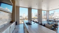 The Sky Lounge in Houston, TX, is an elevated amenity space designed to blend refined interior comfort with sweeping views of the Houston skyline. The entry welcomes guests through steel-framed glass doors set against stone tile flooring, establishing an upscale tone from the start.

Inside, the lounge features distinct seating areas anchored by herringbone-patterned hardwood floors and crisp white walls accented with crown molding. A built-in wet bar and beverage center, framed by a textured stone tile backsplash, serves as a central focal point alongside a wall-mounted television — ideal for gatherings and entertainment. Plush neutral seating arrangements create inviting conversation areas throughout.

Floor-to-ceiling windows and large gridded glass panes flood the interior with natural light while framing unobstructed panoramic views of the Houston skyline. Linen drapery provides an elegant finishing touch to the windowed seating lounge.

The space transitions seamlessly to an open-air terrace through expansive openings, where stone tile flooring, a bar counter with seating, an outdoor television, and lounge ottomans take advantage of the elevated outdoor setting. The terrace offers open-air views of downtown Houston in multiple directions, making it a standout feature of the lounge overall.