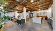 Located at 51 S Coyle St in Pensacola, FL, this thoughtfully designed single-floor space offers a seamless open-concept layout that blends rustic character with modern sophistication. The most striking architectural feature is the reclaimed wood beam ceiling, which runs throughout the entire space, adding warmth and a sense of crafted authenticity overhead. Large black-framed windows flood the interior with natural light, while slate-tiled flooring in cool gray tones grounds the space with a sleek, durable finish.

The layout flows effortlessly from a comfortable lounge area — anchored by a velvet armchair and neutral sofa — into a functional kitchenette and bar zone. This wet bar area is a true standout, featuring stainless steel appliances, rich wood cabinetry, a built-in beverage refrigerator, and a dramatic arabesque-patterned tile accent wall that adds bold visual contrast. A large white quartz island serves as a central hub, illuminated by a pair of geometric brass lantern pendants.

A graceful arched alcove houses an intimate banquette dining nook, adorned with a tropical palm-leaf mural and warm pendant lighting. A stucco fireplace surround and bold abstract wall art add further architectural and artistic interest. The overall ambiance is warm, inviting, and polished — ideal for gathering or entertaining.