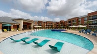 Located at 1502 Partnership Way in Houston, Texas, this single-floor amenity facility offers a well-appointed fitness center and expansive outdoor resort-style pool area. The fitness center is thoughtfully designed with bold, energizing aesthetics, including a vibrant chartreuse accent wall, hexagonal-patterned carpet in shades of yellow, grey, and taupe, and large floor-to-ceiling windows that flood the space with natural light and provide direct views of the pool deck. The gym is equipped with a wide array of strength and cardio machines, including cable systems, rowing machines, recumbent bikes, and treadmills, alongside a dedicated stretching and accessory station with built-in shelving. Mounted flat-screen TVs are positioned throughout for an engaging workout experience. The entry corridor features a built-in water bottle refill station, decorative indoor plants, and curated abstract artwork, contributing to a welcoming and polished atmosphere. Large glass doors provide seamless indoor-outdoor connectivity to the exterior, where vending machines are conveniently located. The outdoor pool area is a standout feature, offering a sprawling resort-style swimming pool with in-water lounge chairs, umbrella-shaded seating, and a connected spa, all framed by the multi-story brick residential buildings surrounding the courtyard.