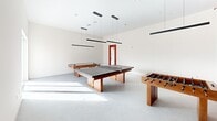 Located at 401 Convery Blvd in Perth Amboy, NJ, this single-floor recreational space is designed to deliver an engaging and energetic environment for leisure and social activity. The game room serves as the centerpiece, boasting a bold terracotta-red accent wall that adds a striking visual contrast to the crisp white walls and neutral concrete-style flooring throughout. The room is generously proportioned, comfortably accommodating a full-size ping pong table, an air hockey table, and a foosball table simultaneously. Overhead, a series of sleek linear pendant lights hang from the elevated ceiling, providing well-distributed illumination across the play area. Large sliding glass doors open directly to an outdoor terrace, flooding the space with abundant natural light and offering views of the surrounding neighborhood. The seamless indoor-outdoor connection enhances the open, airy feel of the room. A dedicated bathroom is thoughtfully appointed with rich brown ceramic wall tiles, a terrazzo-patterned floor, a modern pedestal sink with an oval mirror, and accessible fixtures — combining functionality with a distinctive, design-forward aesthetic that complements the overall character of the space.