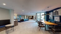 Located at 2200 John Muir Pkwy in Hercules, California, this single-floor community lounge is a thoughtfully designed gathering space that blends contemporary style with comfortable functionality. The expansive open-plan layout features large-format hexagonal tile flooring throughout, creating a cohesive and sophisticated foundation for the various distinct zones within the space.

A standout focal point is the bold fireplace wall, clad in a striking black chevron-patterned tile with gold accents, flanked by a built-in wooden bookshelf and a decorative log storage display. Adjacent to this feature wall, a cozy seating area is anchored by a rich red area rug and leather accent chairs, lending warmth to the space.

The central lounge area offers generous seating with a teal sofa, upholstered armchairs, and a dark area rug, centered around a wall-mounted TV and curated wall art. A built-in wet bar with under-counter refrigeration and marble-look countertops lines one wall, ideal for social gatherings.

A dedicated dining zone features a long communal wood table beneath industrial pendant lights, set against a deep navy accent wall adorned with a curated gallery of artwork. A banquette seating nook with navy cushions and wire-frame side tables provides an additional casual setting. Floor-to-ceiling glass doors open to an outdoor courtyard, filling the space with natural light and greenery views.